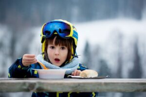 Kind met skihelm en skipak eet soep aan een tafeltje op de skipiste